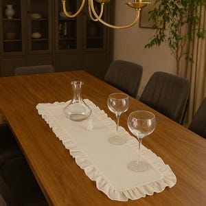 May include: A wooden dining table set for a meal. A cream-colored ruffled table runner holds a clear glass decanter, two wine glasses, and a small decorative object. A gold chandelier hangs above.