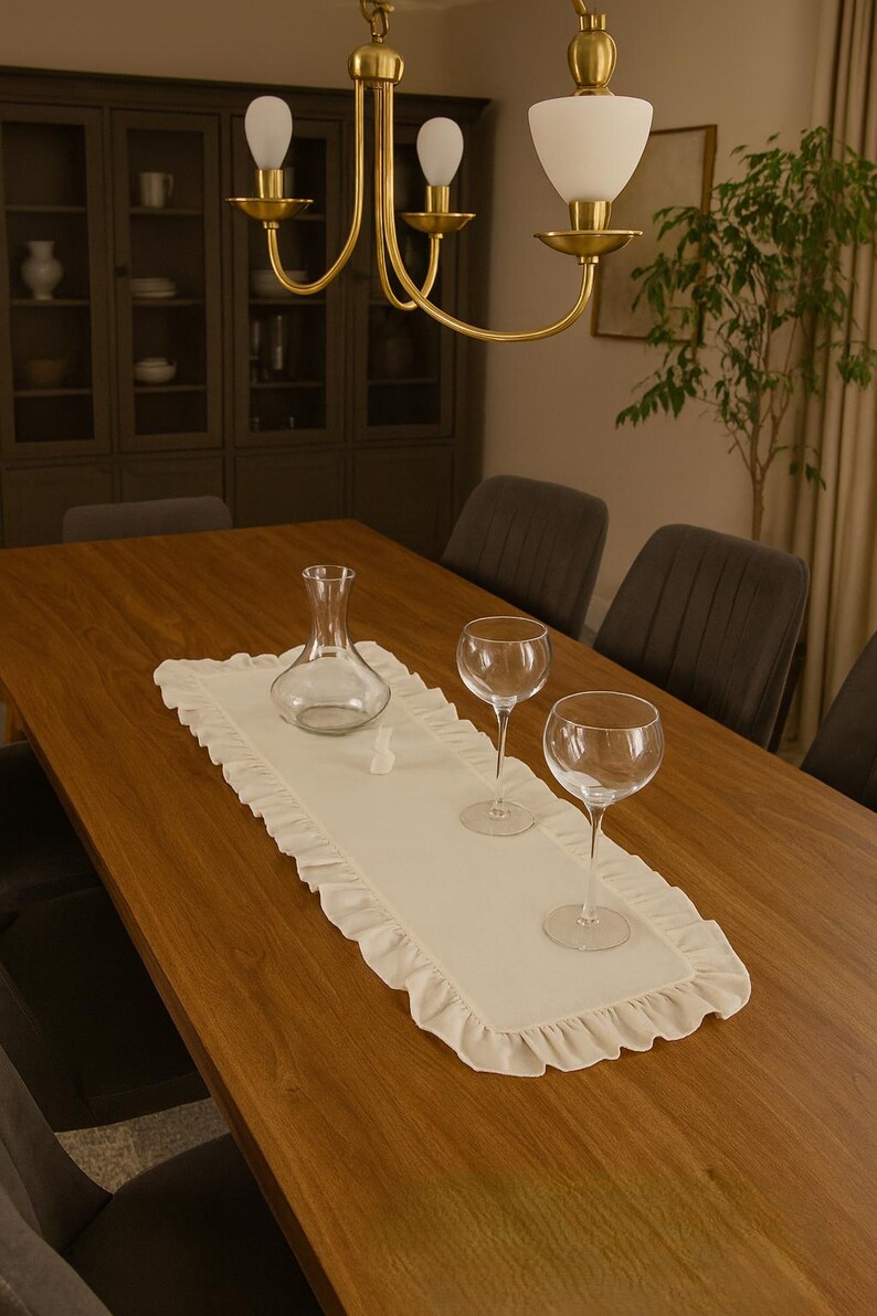 May include: A wooden dining table set for a meal. A cream-colored ruffled table runner holds a clear glass decanter, two wine glasses, and a small decorative object. A gold chandelier hangs above.