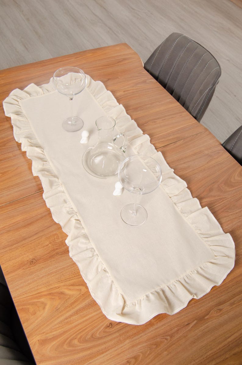 May include: A cream-colored table runner with ruffled edges, set on a wooden table. Two wine glasses, a carafe, and decorative pieces are arranged on the runner, suggesting a dining or event setting.