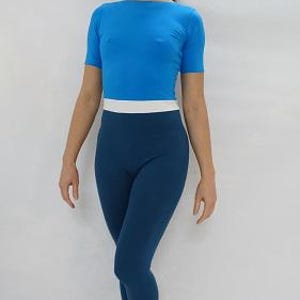 Op de afbeelding: Een vrouw draagt een tweedelige sportoutfit. De top is een felblauw shirt met korte mouwen. De onderkant is een donkerblauwe legging met een witte band in de taille. De outfit is nauwsluitend en lijkt gemaakt van een rekbaar materiaal.