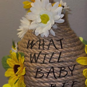 May include: A hanging, beehive-shaped ornament made of coiled rope, decorated with yellow sunflowers and white daisies. The text "WHAT WILL BABY BEE" is written on the front. A charming piece for a baby shower or nursery.