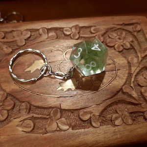 Pode incluir: Um porta-chaves com um dado de vinte faces translúcido, de cor verde-clara, feito de resina. O dado contém números brancos e está preso a uma argola de metal prateado por uma corrente curta. Um segundo dado é parcialmente visível.