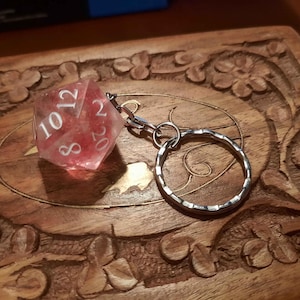 May include: A translucent pink twenty-sided die keyring featuring white numbers. The die is attached to a silver-toned metal split ring by a small jump ring and a short chain link, creating a functional accessory for keys or bags.
