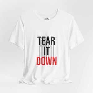 Puede incluir: Camiseta blanca con el texto "TEAR IT DOWN" en negro y rojo, con un diseño estilizado de la bandera estadounidense. La camiseta está hecha de un material suave y ligero.