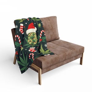May include: A dark green throw blanket draped over a brown chair. The blanket features a festive design with a cannabis bud wearing a Santa hat, candy canes, holly, and snowflakes. The blanket is made of a soft material.