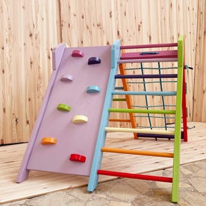 May include: A colorful wooden climbing frame with a slide and a rope ladder. The slide is purple with colorful handholds and the ladder has rainbow colored rungs.