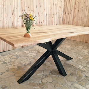 Peut inclure: Table rectangulaire en bois avec un bord naturel et un piètement en métal noir. La table est en bois clair et a un bord brut sur un côté. Un petit vase avec des fleurs séchées est posé sur la table.