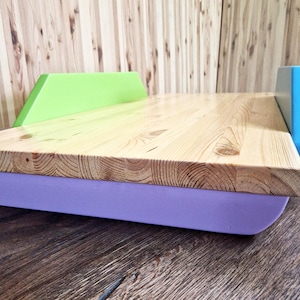 Puede incluir: Un escritorio portátil de madera con una superficie de madera clara, una base lavanda y detalles en verde y azul. El escritorio está diseñado con un borde elevado y una base curva.