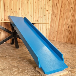 Può includere: Uno scivolo in legno blu brillante, appoggiato su una base in legno naturale. Lo scivolo è inclinato verso il basso, con bordi rialzati. Lo sfondo presenta una parete in legno e un tavolo con gambe nere. Lunghezza di circa 120 cm.