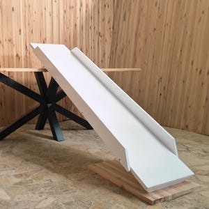 Könnte beinhalten: Eine weiße Holzrutsche mit erhöhten Seiten, auf einer Basis aus Naturholz. Die Rutsche ist abgewinkelt, was auf ein verspieltes Design hindeutet. Im Hintergrund ein Tisch mit schwarzem Metallgestell.