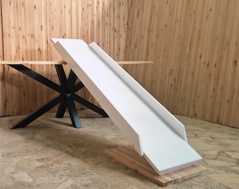 May include: A white wooden slide with raised sides, set on a natural wood base. The slide is angled, suggesting a playful design. A table with a black metal base is in the background.