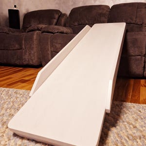 May include: A white wooden pet ramp with a gently sloped design. The ramp is designed to assist small dogs or cats in reaching elevated surfaces. The ramp is placed on a patterned rug, with a brown sofa in the background.