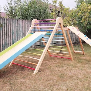 May include: A wooden outdoor playset with a rainbow-colored ladder, a slide, and a climbing wall. The playset is made of natural wood and has a light blue and green slide.