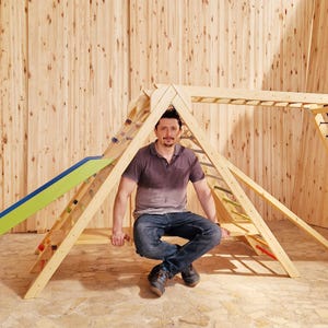 Może przedstawiać: Drewniana konstrukcja zabawowa ze zjeżdżalnią, ścianką wspinaczkową i drabiną linową. Osoba siedzi na platformie konstrukcji.