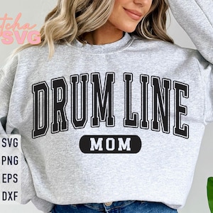 Pode incluir: Camisola cinza claro com as palavras "DRUM LINE MOM" em letras pretas. O texto é arqueado sobre o peito. A camisola também tem as palavras "SVG, PNG, EPS, DXF" numa pilha vertical no lado esquerdo.