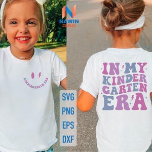 Puede incluir: Una camiseta blanca con el texto "IN MY KINDERGARTEN ERA" en morado, azul y rosa en la espalda. La parte delantera presenta una carita sonriente y las palabras "KINDERGARTEN ERA" en rosa. También hay un gráfico de rayo.