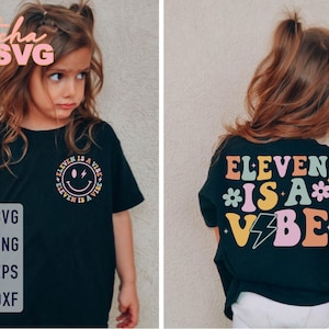 Op de afbeelding: Zwart T-shirt met de tekst "ELEVEN IS A VIBE" in een kleurrijk retro lettertype. De voorkant van het shirt heeft een smiley met de tekst "ELEVEN IS A VIBE" eromheen. Het shirt wordt gedragen door een jong persoon.