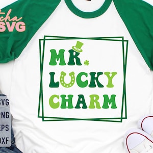 Puede incluir: Camiseta raglán blanca y verde con el texto "MR. LUCKY CHARM" en tonos verdes. El diseño incluye un sombrero de duende y una herradura. La camiseta tiene mangas verdes y cuello verde. Un tema del Día de San Patricio.
