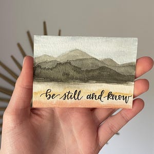 May include: Watercolor painting of a mountain range with a light blue sky and a peach colored sunset. The text "be still and know" is written in black ink at the bottom of the painting.