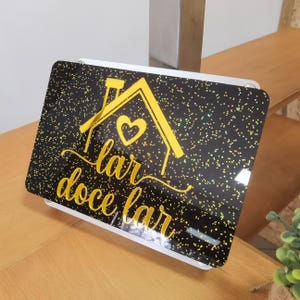 May include: A decorative sign with a black background speckled with glitter. A yellow house outline with a heart and the words "lar doce lar" are featured on the sign. The sign is mounted on a white base.