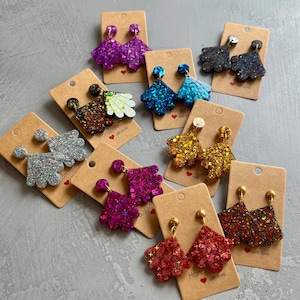 Pode incluir: Conjunto de brincos feitos à mão com glitter e acabamentos iridescentes. Os brincos são exibidos em cartão castanho com o texto "Handmade with love". Os brincos são de várias cores, incluindo prata, ouro, azul, vermelho, roxo e preto.