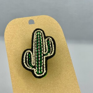 Peut inclure: Un patch brodé de cactus vert et or sur une carte marron avec le mot "Charly" écrit dessus.