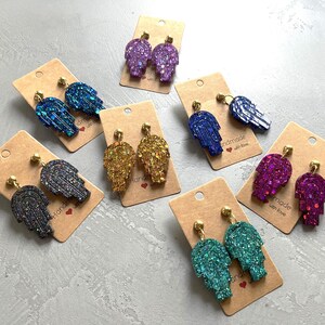 Peut inclure: Assortiment de boucles d'oreilles en paillettes faites à la main dans différentes couleurs, notamment le violet, le bleu, le vert, l'or et le noir. Chaque paire présente un motif de main de hamsa. Les boucles d'oreilles sont présentées sur des cartes avec le texte "Handmade with love".