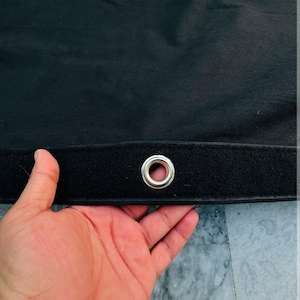 Puede incluir: Tela negra con un ojal plateado. La tela tiene una superficie texturizada y una tira de velcro negra. El ojal está centrado en la tira de velcro. La imagen muestra una mano sosteniendo la tela.