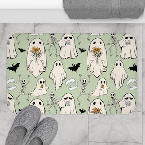 May include: A rectangular bath mat with a light green background featuring a repeating pattern of cartoon ghosts, skeletons, bats, and vampire teeth. Some ghosts hold flowers. The mat is next to gray slippers.