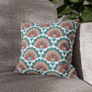 Könnte beinhalten: Quadratisches Zierkissen mit sich wiederholendem Blumenmuster. Das Design zeigt stilisierte orangefarbene Blumen mit türkisfarbenen und weißen Akzenten auf weißem Hintergrund. Das Kissen liegt auf einem grauen Polstersofa.