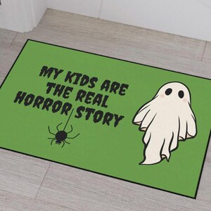 以下が含まれることがあります： 「MY KIDS ARE THE REAL HORROR STORY」の文字が入った緑色の長方形のドアマット。漫画のゴーストとクモもマットに描かれています。マットには黒い縁取りがあります。
