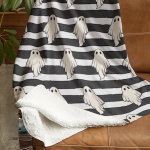 May include: A Halloween-themed blanket featuring a pattern of friendly ghost figures on a black and white striped background. The blanket is draped over a brown leather couch, with a soft, white sherpa lining visible.