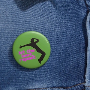 May include: A round, green button with a black silhouette of a person dancing. The button has the text "hit the beat, repeat" in pink. The button is set against a blue denim background.