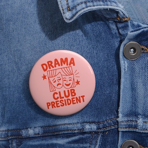 Può includere: Un bottone rosa chiaro con la scritta "DRAMA CLUB PRESIDENT" in rosso. Il bottone presenta una grafica di due maschere teatrali, una sorridente e l'altra accigliata, con stelle. Il bottone è appuntato su una giacca di jeans blu.
