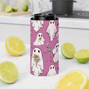 May include: A pink can cooler featuring a pattern of white ghosts and skeletons. Some ghosts hold flowers. The cooler is designed to keep beverages cold and is surrounded by lemons and limes.