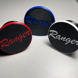 May include: Three round, black accessories with the word "Ranger" in different colors: red, blue, and white. Each has a textured surface and a colored rim. The accessories are displayed on a white surface.