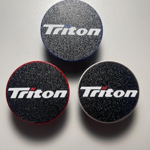 May include: Three circular automotive accessories with the word "Triton" in white. One has a blue border, one a red border, and the third has a silver border. All have a black textured surface.