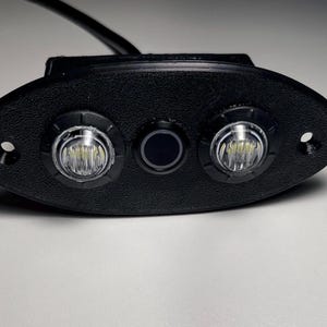 May include: A black oval-shaped light fixture with two white LED lights and a black button in the center.