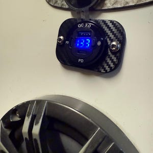 Może przedstawiać: Czarna ładowarka USB QC 3.0 z wzorem z włókna węglowego. Ładowarka wyświetla niebieski cyfrowy odczyt 13,3. Ładowarka jest zamontowana na białej powierzchni z innymi widocznymi elementami.