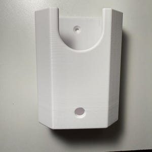 May include: A white, rectangular wall-mounted holder with a curved top and a circular hole at the bottom. The holder has a semi-circular cutout at the top and a small hole near the top.