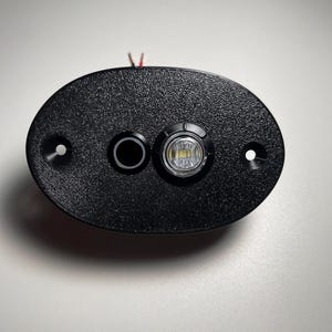 Pode incluir: Uma caixa de plástico preto com uma luz LED branca redonda e um botão preto redondo. A caixa tem dois pequenos orifícios de cada lado.