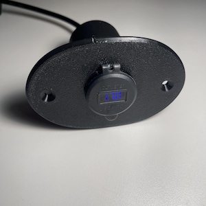 May include: A black oval-shaped electrical outlet with a blue digital display showing "12.7V". The outlet is mounted on a black surface.