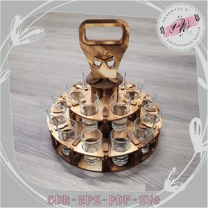 Puede incluir: Un soporte de madera para vasos de chupito con asa, diseñado para contener varios vasos. El soporte tiene tres niveles circulares, cada uno con ranuras para los vasos. La parte superior presenta un asa y un recorte decorativo de dos vasos brindando. El texto "CDR - EPS - PDF - SVG" está en la parte inferior.