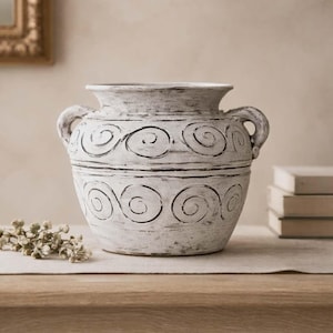 May include: A white ceramic vase with black spiral patterns and two handles. The vase has a distressed finish, giving it a vintage appearance. It sits on a wooden table, suggesting a home decor theme. The vase is approximately 25 cm tall.