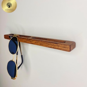 May include: A wooden sunglasses holder with a dark stain, featuring three recessed slots and a hanging pair of sunglasses. The sunglasses have gold frames and blue lenses. The holder is mounted on a white wall. The logo "Tree Bees" is visible.