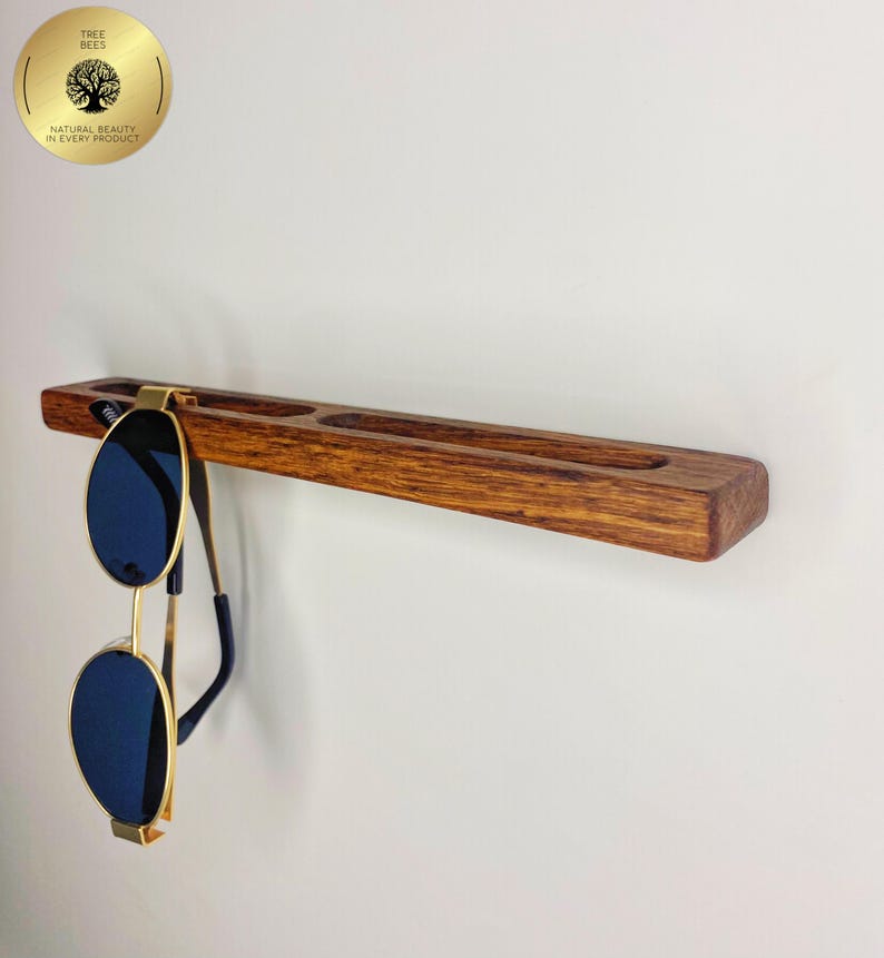 May include: A wooden sunglasses holder with a dark stain, featuring three recessed slots and a hanging pair of sunglasses. The sunglasses have gold frames and blue lenses. The holder is mounted on a white wall. The logo "Tree Bees" is visible.