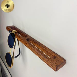 May include: A wooden sunglasses holder with a dark brown finish, mounted on a white wall. The holder has slots for multiple pairs of glasses and is holding a pair of gold-rimmed sunglasses with black lenses. The holder is approximately 30 cm long.