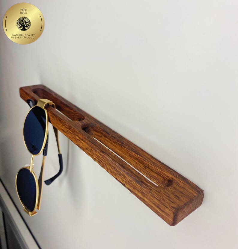 May include: A wooden sunglasses holder with a dark brown finish, mounted on a white wall. The holder has slots for multiple pairs of glasses and is holding a pair of gold-rimmed sunglasses with black lenses. The holder is approximately 30 cm long.