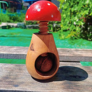 May include: A wooden nutcracker shaped like a mushroom. The mushroom cap is painted red with white spots. The nutcracker has a wooden screw mechanism and a round opening to hold nuts. A walnut is visible inside.