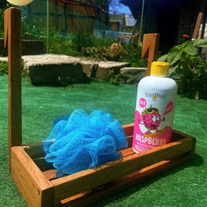 Puede incluir: Un organizador de ducha de madera con una esponja de malla azul y una botella de gel de ducha con aroma a frambuesa. El organizador es de madera marrón claro y tiene un asa para colgar. La botella de gel de ducha es blanca con una etiqueta rosa.